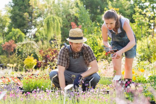 In a vibrant, well-maintained garden in Hampstead, two women are engaged in gardening activities amidst a variety of colorful flowering plants and lush greenery. The woman on the left, wearing a straw hat and a checkered shirt with gardening gloves, is kneeling close to the ground, carefully tending to a garden bed filled with pink and purple flowers. The woman on the right, dressed in denim overalls and a sleeveless top, is leaning forward, holding pruning shears and inspecting a plant or flower. The background features a mix of dense trees, including a weeping willow and tall shrubs, providing a natural, leafy backdrop. The garden surface includes neatly edged flower beds, alongside a grassy lawn area with small patches of soil and stepping stones visible. Sunlight illuminates the scene, indicating clear weather, and the environment suggests a well-organized outdoor space suitable for ongoing gardening and lawn care, reflective of services by Landscapers Hampstead on the postcode area around NW3. This natural and peaceful setting demonstrates outdoor maintenance efforts connected to local gardening services in Hampstead's residential gardens.