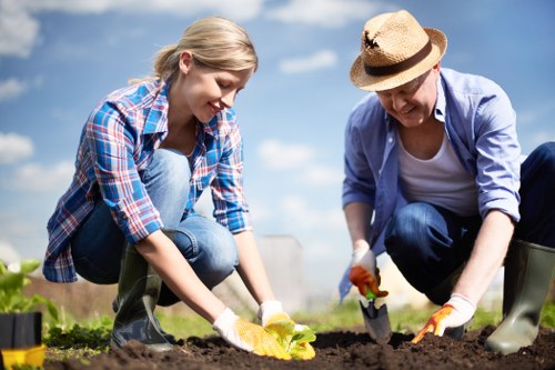 Two individuals, a young woman and an older woman, are gardening outdoors in a well-maintained backyard with clear weather. The young woman, dressed in a blue plaid shirt, jeans, and rubber boots, is kneeling on the soil, planting or tending to a small yellow flowering plant. The older woman, wearing a light blue shirt, dark trousers, a wide-brimmed straw hat, and gardening gloves, is crouching beside her, using a trowel to work the soil around the plant. The garden features a lush green lawn with neatly edged flower beds bordered by soil and a paved pathway in the background, with several trees providing shade and a blue sky with some clouds overhead. The scene suggests a focus on outdoor gardening and lawn maintenance, supported by the professional services offered by Landscapers Hampstead in the Hampstead area, as part of their gardening and landscaping offerings.