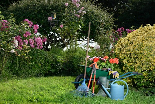 The image depicts a well-maintained garden with a lush, green lawn in the foreground, bordered by various flowering bushes and shrubs, including pink and purple blossoms. In the middle ground, there is a collection of gardening tools and supplies placed on the grass, including a blue watering can, a garden rake, and a small wheelbarrow. Behind these tools, a tall wooden stake supports a flowering rose bush with light pink blossoms. The background features dense foliage and trees, providing a natural backdrop for the landscaped outdoor space. The scene appears to be on a bright, clear day, with natural sunlight illuminating the vibrant plant colors and the tidy appearance of the garden. This outdoor environment exemplifies typical garden maintenance and landscape care, suitable for outdoor gardening services provided by companies like Landscapers Hampstead, contributing to an attractive, well-kept front or back garden in Hampstead, London.