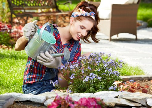 A young woman with a cheerful expression is kneeling in a well-maintained garden in Hampstead, tending to a flowering shrub with small purple and light blue blossoms. She is wearing a plaid shirt, denim jeans, gardening gloves, and a colorful headband, and is holding a silver watering can, watering the plant carefully. The garden features a neatly edged flower bed bordered with flat stones, with visible lush green grass and a variety of plants, including bushes and a small tree in the background. Behind her, there is outdoor furniture with wooden chairs and a paved patio area, suggesting a pleasant outdoor space for relaxation. The scene is brightly lit with natural daylight, indicating a clear, sunny day. This garden setting exemplifies professional landscaping and gardening care, similar to services offered by Landscapers Hampstead, with a focus on healthy plants, tidy surfaces, and a welcoming environment in the Hampstead area, postcode NW3.