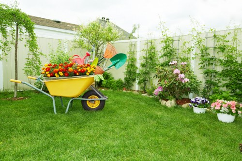A well-maintained backyard garden featuring a lush, green lawn with evenly trimmed grass in the foreground, bordered by a white fence behind which a variety of climbing plants and greenery grow along the top. To the right, there are colorful flower pots with blooming flowers, including pink and purple blossoms, arranged along the edge of the garden bed. In the middle, a yellow wheelbarrow filled with bright red, orange, and yellow flowers is positioned on the grass, with garden implements such as a green watering can and an orange umbrella nestled beside it. The background shows small trees and shrubs providing natural privacy and shade, with an overcast sky overhead. The overall scene reflects a tidy, inviting outdoor space suitable for gardening activities and outdoor relaxation, maintained by local landscaping services like Landscapers Hampstead, serving the Hampstead area and postcode NW3.