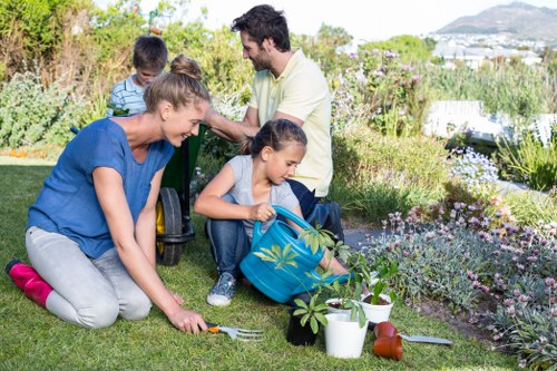 A family gardening outdoors on a neatly maintained lawn in Hampstead, with a woman planting a small shrub in the foreground, surrounded by gardening tools and potted plants. In the background, a man and a young girl are engaged with additional garden plants, with the girl holding a blue watering can. The garden features a lush, green grass area, edged by flower beds with blooming flowers and foliage. There are visible garden tools such as a small rake and watering cans, and the overall scene indicates active garden maintenance in a peaceful outdoor space typical of Hampstead's residential gardens. Bright daylight illuminates the scene, emphasizing natural tones of green, brown, and floral colours, suggestive of good weather suitable for outdoor gardening activities. This setting reflects professional gardening practices and outdoor upkeep services offered in the Hampstead area by Landscapers Hampstead, with a focus on garden aesthetics and horticultural care.