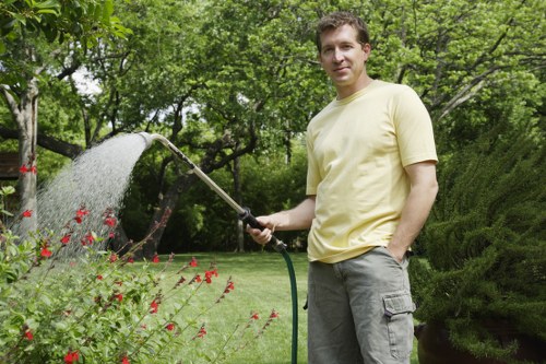 A man in a light yellow t-shirt and grey trousers standing in a well-maintained garden with lush green trees and shrubs in the background. He is holding a garden hose and watering a flower bed with red blooms and green foliage. The lawn area adjacent to the flower bed has dense, evenly cut grass. The scene is outdoors on a clear, possibly sunny day with natural light illuminating the garden. The garden features a variety of plant textures, with soft grass, flowering plants, and shrubbery, typical of landscaped outdoor spaces in Hampstead. This setting reflects professional gardening and lawn care services, with a focus on healthy plant growth and neat, attractive outdoor environments, consistent with offerings from Landscapers Hampstead.