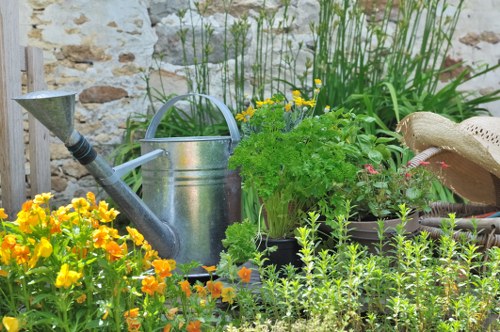 A small, well-maintained garden features a variety of plants and gardening tools arranged against a rustic stone wall. In the foreground, bright yellow and orange flowering plants fill the space with vibrant colour, contrasting with the lush green foliage of surrounding shrubs and flowers. Behind these, a light grey watering can sits atop the soil, with a weathered, straw hat resting nearby on a stone surface. Tall, slender green grasses and leafy bushes occupy the background, creating a sense of layered planting typical of a landscaped outdoor space in Hampstead. The lighting suggests a partly cloudy day, with natural daylight illuminating the textures of leaves, flowers, and gardening equipment. This scene exemplifies a well-kept garden environment suitable for professional landscaping and gardening services offered by companies like Landscapers Hampstead, emphasizing outdoor care, planting, and garden maintenance in a UK residential setting.