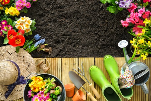 The image depicts a small, neatly maintained garden area with a section of dark, freshly turned soil bordered by a wooden decking. In the foreground, various gardening tools and accessories are arranged, including bright green rain boots, a watering can, a gardening trowel, and small plant pots. Adjacent to these items, there is a woven straw sunhat with a purple ribbon, along with a black container filled with a colourful assortment of flowers such as primroses and pansies, highlighting seasonal planting efforts. The garden features a vibrant flower border with multicoloured blooms like pink, purple, yellow, and red flowers, which frame the edge of the prepared soil. In the background, lush green hedges and small trees provide privacy and natural beauty to the outdoor space. The overall scene suggests a well-organised garden that is actively maintained, with seasonal planting and soil preparation, typical of professional gardening services provided by Landscapers Hampstead in NW London, supporting sustainable planting and outdoor care.