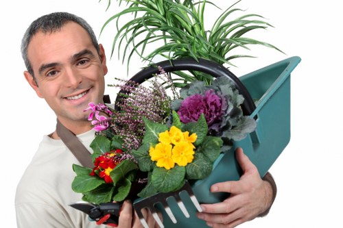 A smiling man with short dark hair and a light-colored shirt is holding a green garden watering can filled with a variety of colourful plants, including purple, yellow, and red flowers, as well as green foliage. The watering can is positioned over a black pot, and the man appears to be presenting the plants, suggesting a focus on gardening or plant care. The background is plain and white, emphasizing the gardening theme and visually connecting it to outdoor landscape maintenance and garden planting services relevant to Hampstead and surrounding areas. The scene conveys a professional and approachable outdoor gardening service environment, highlighting plant selection, garden beautification, and the nurturing of healthy greenery.