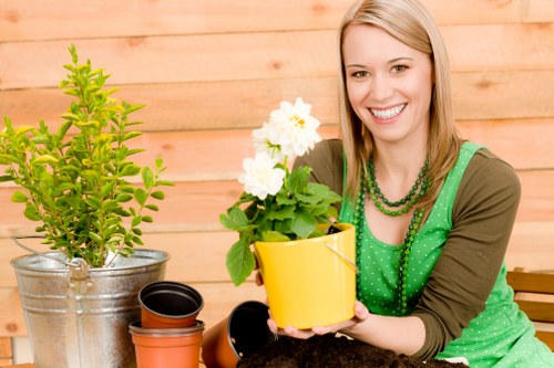 A woman with blonde hair and a bright smile is sitting in a garden patio area, holding a yellow flower pot containing a white flowering plant with lush green leaves. To her left, there is a larger metal pot with a green shrub or small tree, and nearby are empty smaller plant pots made of terracotta and black plastic. The background features a wooden fence, typical of a suburban or residential garden in Hampstead, with a warm, natural tone that complements the outdoor setting. The surface she is sitting on appears to be part of a garden table, possibly made of wood or metal, and the scene suggests an organized, well-maintained outdoor space designed for gardening activities or enjoying nature. The overall atmosphere is bright and inviting, with natural daylight illuminating the scene, enhancing the vivid green of the plants and the cheerful expression of the woman. This image connects to general gardening services offered by Landscapers Hampstead, especially those related to planting, container gardening, and outdoor space enhancement in the Hampstead area, postcode NW3, suitable for clients seeking professional lawn care, garden design, or plant maintenance.