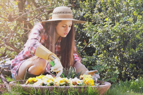A young woman wearing a wide-brimmed straw hat, plaid shirt, and garden gloves is kneeling on a well-maintained lawn in a garden, tending to a flower bed filled with vibrant yellow and white flowers. She appears to be planting or weeding, with her focus directed at the blooms. The garden features lush green hedges and dense foliage in the background, with sunlight filtering through the leaves, creating a warm and inviting outdoor environment. The area surrounding her includes a patch of grass, garden soil, and possible paving or stone edging, indicative of landscaped outdoor space typical of Hampstead gardens. The scene reflects attentive gardening activity that a professional landscaper or gardening service, such as Landscapers Hampstead, might support through detailed garden care, lawn maintenance, and planting services. The overall setting emphasizes a cultivated and natural outdoor environment, ideal for enhancing local gardens and outdoor spaces in the Hampstead area.