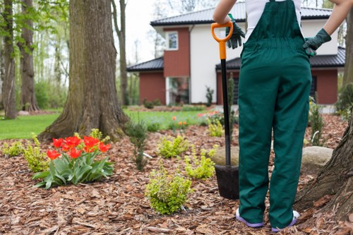 A person dressed in dark green gardening overalls is standing in a landscaped front garden, holding a small shovel with an orange handle. The garden features a neatly mulched flower bed with vibrant red tulips and various small green shrubs. In the background, there are mature trees with thick trunks and a modern, red-brick house with white window frames and a dark tiled roof, situated in Hampstead. The lawn area appears well-maintained with lush, green grass, and the garden borders are clearly defined with natural stone edging. The scene suggests a bright, mild day with soft natural light illuminating the outdoor space, captured in a natural outdoor setting typical of a residential garden in London, aligning with professional gardening and landscaping services offered by Landscapers Hampstead. The overall environment combines natural garden textures, seasonal flower colours, and structured landscaping features, reflecting a healthy, well-kept outdoor space ready for ongoing maintenance or improvements.